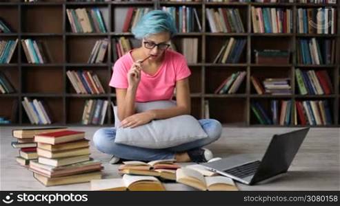 Concentrated college female student sitting on the floor, surronded by heap of books, researching with and laptop in university library. Serious hipster girl with blue hair and trendy eyewear studying in library with laptop on the internet.