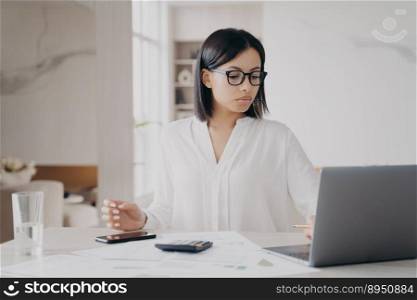 Concentrated european businesswoman in glasses and white blouse is working remote from home. Manager, accountant or business assistant is doing paperwork using laptop. Success and career concept.. Concentrated european businesswoman is working remote from home. Success and career concept.
