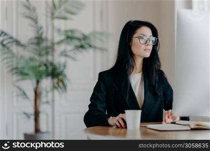 Concentrated female entrepreneur wears elegant black costume, works at computer, sits at table in cozy work place wears transparent glasses browses internet makes business project thinks over strategy