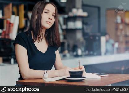 Concentrated female journalist thinks about new article, writes down information into notepad, drinks hot aromatic beverage in coffee shop, modern smart phone on table near. Working process.