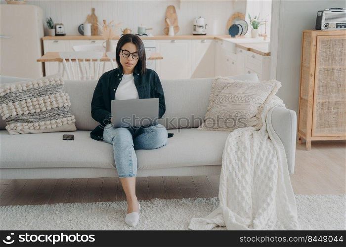 Concentrated girl typing on computer. Hispanic business woman is working on startup project from home. Lady freelancer is sitting in couch with laptop. Comfortable workplace in modern apartment.. Concentrated girl typing on laptop, working from home. Comfortable workplace in modern apartment.