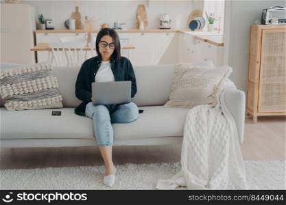 Concentrated girl typing on computer. Hispanic business woman is working on startup project from home. Lady freelancer is sitting in couch with laptop. Comfortable workplace in modern apartment.. Concentrated girl typing on laptop, working from home. Comfortable workplace in modern apartment.