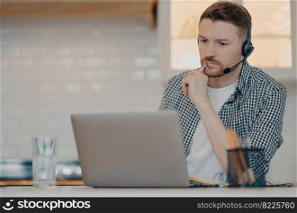 Concentrated guy looking at laptop screen and holding pen while having video call with colleagues or surfing internet, male freelancer working remotely at home, being serious and focused at work. Young bearded man using headset and laptop while working at home