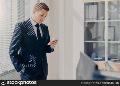 Concentrated male boss, keeps hand in pocket, focused into screen of smart phone, searches useful information for new startup, poses in office, dressed elegantly. Man chats on cellular at cabinet