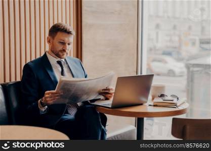 Concentrated male business company worker in blue formal suit sitting in corner of cafe next to big window, reading newspaper closely, open work laptop and coffee in front of him on table. Male business company worker reading newspaper while working in cafe