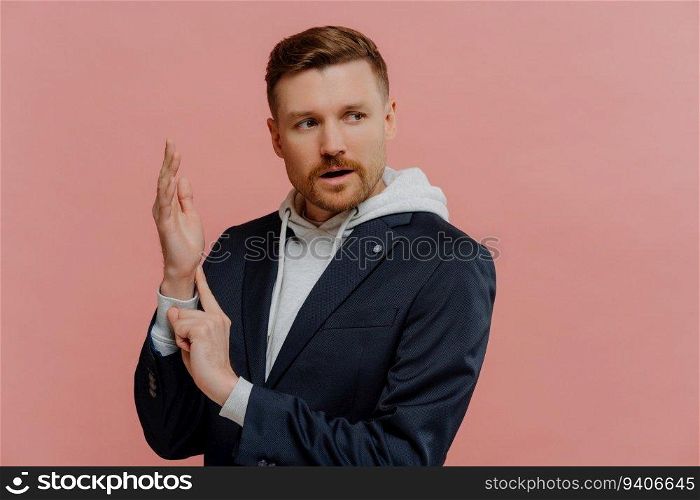 Concentrated red-haired man measuring pulse, looking aside. Casual fashion, pastel pink background.