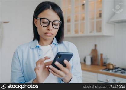 Concentrated serious woman is surfing through internet with mobile phone. Remote worker in glasses texting and consulting online or using application. Distant work on quarantine from home.. Concentrated woman texting and consulting online or using application. Distant work on quarantine.