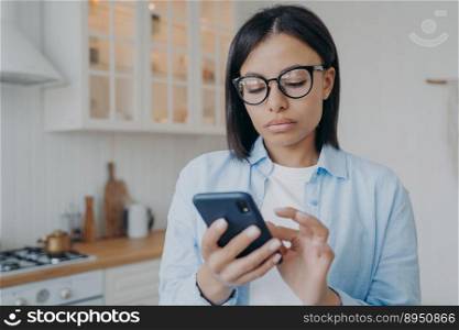 Concentrated serious woman is surfing through internet with mobile phone. Remote worker in glasses texting and consulting online or using application. Distant work on quarantine from home.. Concentrated woman texting and consulting online or using application. Distant work on quarantine.