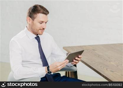 Concentrated sideways shot of businessman in formal clothes holds modern tablet computer, checks data or email box, connected to wireless internet, sits in modern spacious cabinet. Business concept