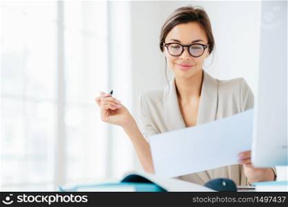 Concentrated successful businesswoman looks attentively in paper, studies terms of contract, holds pen, writes in documentations, dressed formally, poses at desktop against white spacious interior