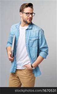 Concentrated young bearded man wearing glasses dressed in jeans shirt holding laptop isolated over grey studio background.. Concentrated young bearded man wearing glasses dressed in jeans shirt holding laptop isolated over grey studio background