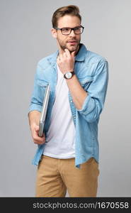 Concentrated young bearded man wearing glasses dressed in jeans shirt holding laptop isolated over grey studio background.. Concentrated young bearded man wearing glasses dressed in jeans shirt holding laptop isolated over grey studio background