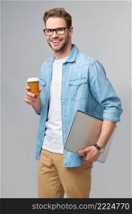 Concentrated young bearded man wearing glasses dressed in jeans shirt holding laptop and cup of coffee to go isolated over grey studio background.. Concentrated young bearded man wearing glasses dressed in jeans shirt holding laptop and cup of coffee to go isolated over grey studio background