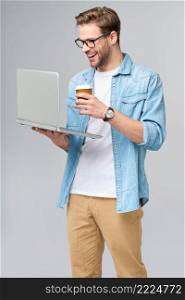 Concentrated young bearded man wearing glasses dressed in jeans shirt holding laptop and cup of coffee to go isolated over grey studio background.. Concentrated young bearded man wearing glasses dressed in jeans shirt holding laptop and cup of coffee to go isolated over grey studio background
