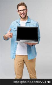 Concentrated young bearded man wearing glasses dressed in jeans shirt holding laptop isolated over grey studio background.. Concentrated young bearded man wearing glasses dressed in jeans shirt holding laptop isolated over grey studio background