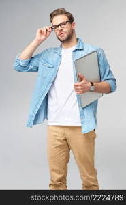 Concentrated young bearded man wearing glasses dressed in jeans shirt holding laptop isolated over grey studio background.. Concentrated young bearded man wearing glasses dressed in jeans shirt holding laptop isolated over grey studio background