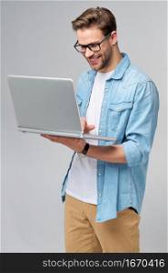 Concentrated young bearded man wearing glasses dressed in jeans shirt using laptop isolated over grey studio background.. Concentrated young bearded man wearing glasses dressed in jeans shirt using laptop isolated over grey studio background