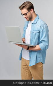 Concentrated young bearded man wearing glasses dressed in jeans shirt using laptop isolated over grey studio background.. Concentrated young bearded man wearing glasses dressed in jeans shirt using laptop isolated over grey studio background