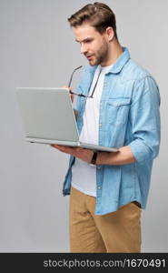 Concentrated young bearded man wearing glasses dressed in jeans shirt using laptop isolated over grey studio background.. Concentrated young bearded man wearing glasses dressed in jeans shirt using laptop isolated over grey studio background