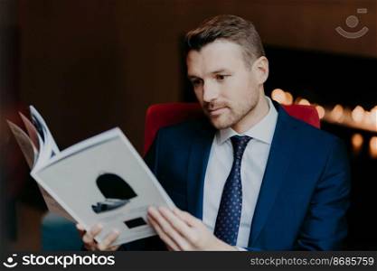 Concentrated young boss has stubble, being well dressed, reads interesting journal while sits at cafe and waits for order, reads interesting information of business sphere. Male employee with magazine
