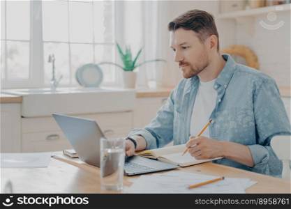 Concentrated young businessman writing notes on agenda and working remotely on laptop in internet from home while sitting at kitchen table. Freelance and work online concept. Concentrated young businessman writing notes on agenda and working remotely on laptop