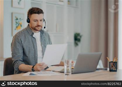 Concentrated young guy sitting in living room with headset and using laptop while holding document and looking on it, male freelancer analyzing business data. Remote work at home. Young man in plaid shirt working from home