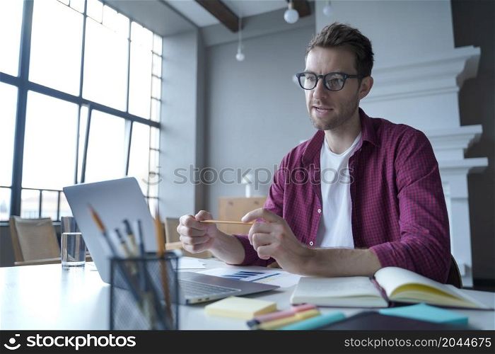 Concentrated young man private financial advisor conducts training courses for individuals or companies online, sitting in front of laptop at modern home office with large windows on background. Focused man private financial advisor conducts training courses for individuals or companies online