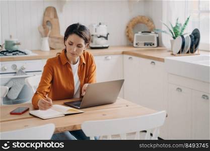 Concentrated young spanish woman is entrepreneur and remote worker. Freelance and distance work concept. Kitchen interior, worktops and cuisines. Daylight through window.. Concentrated young woman is entrepreneur and remote worker. Freelance and distance work concept.