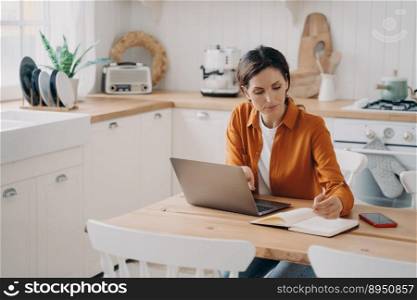 Concentrated young spanish woman is entrepreneur and remote worker. Freelance and distance work concept. Kitchen interior, worktops and cuisines. Daylight through window.. Concentrated young woman is entrepreneur and remote worker. Freelance and distance work concept.