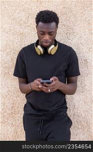 Concentrated young stylish African American man with dark curly hair in casual clothes and headphones on neck standing on street near beige concrete wall and messaging on mobile phone. Serious black male using smartphone near beige wall on street