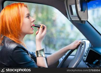 Concept of danger driving. Young woman driver red haired teenage girl painting her lips doing make up while driving the car.