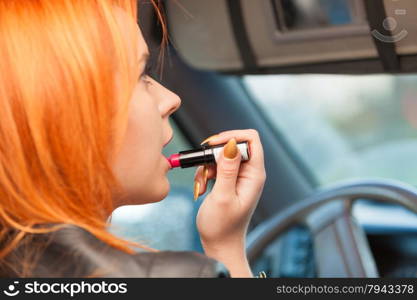 Concept of danger driving. Young woman driver red haired teenage girl painting her lips doing applying make up while driving the car.