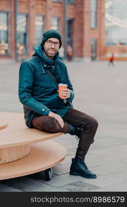 Concept of drinks and lifestyle. Carefree satisfied European man with bristle enjoys sunny day and hot beverage, smiles happily, sits against street background. Outdoor shot of handsome hipster