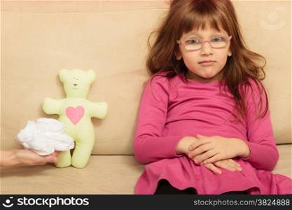 Concept of pregnancy, family, issue, childhood. Sad thoughtful little girl sitting on sofa at home thinking on not wanting sibling. Teddy bear toy, small shoes for unborn baby lying beside