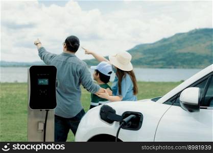 Concept of progressive happy family enjoying their time at green field and lake with electric vehicle. Electric vehicle driven by clean renewable from eco-friendly power sauce.. Concept of progressive happy family at green field, lake with electric vehicle.