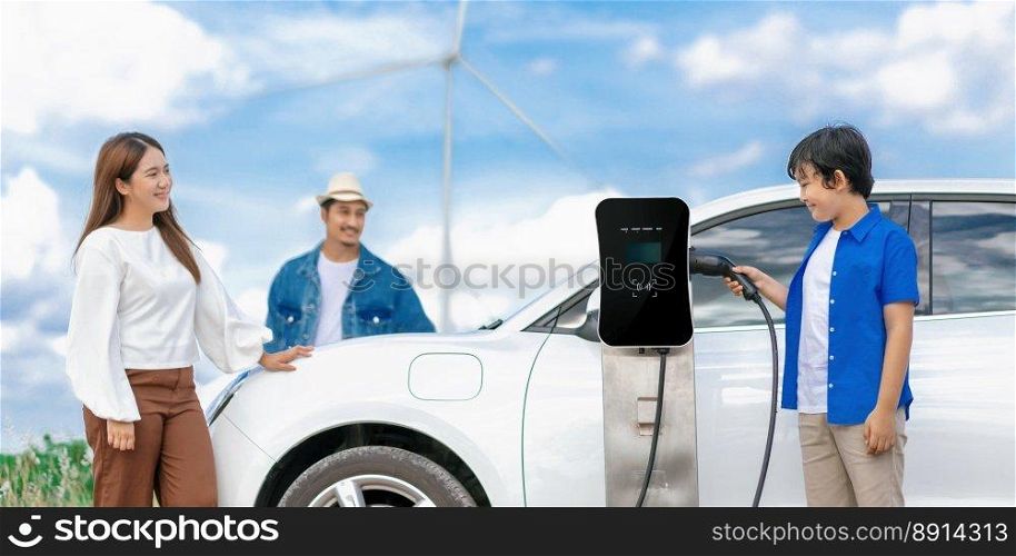 Concept of progressive happy family enjoying their time at wind farm with electric vehicle. Electric vehicle driven by clean renewable energy from wind turbine generator for charging station.. Concept of progressive happy family at wind farm with electric vehicle.