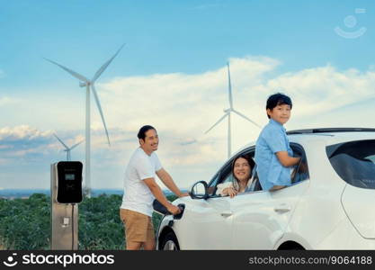 Concept of progressive happy family enjoying their time at wind farm with electric vehicle. Electric vehicle driven by clean renewable energy from wind turbine generator for charging station.. Concept of progressive happy family at wind farm with electric vehicle.