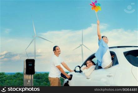 Concept of progressive happy family holding windmill toy and relax at wind farm with electric vehicle. Electric vehicle driven by clean renewable energy from wind turbine generator to charger station.. Concept of progressive happy family at wind turbine with electric vehicle.