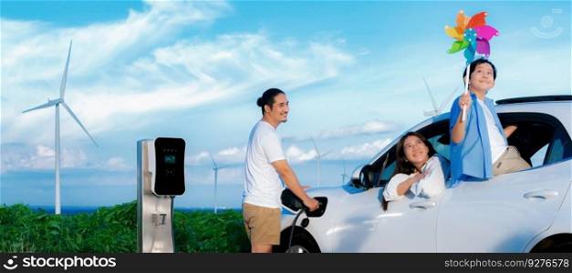 Concept of progressive happy family holding windmill toy and relax at wind farm with electric vehicle. Electric vehicle driven by clean renewable energy from wind turbine generator to charger station.. Concept of progressive happy family at wind turbine with electric vehicle.
