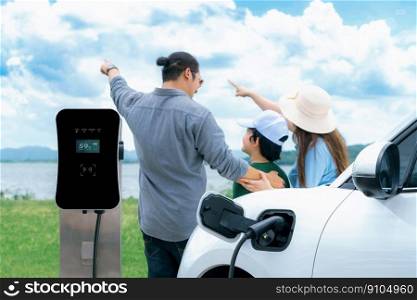 Concept of progressive happy family with electric vehicle enjoy their time at natural outdoor scenic with lake, greenfield and cloudscape background. Electric vehicle driven by green renewable energy.. Concept of progressive happy family with EV car at natural outdoor scenic.