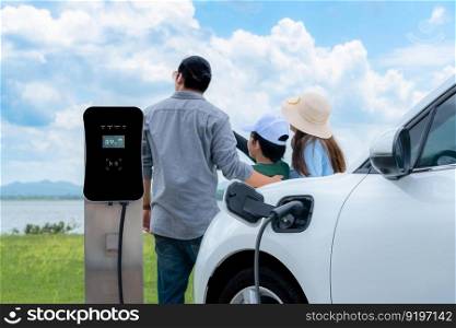 Concept of progressive happy family with electric vehicle enjoy their time at natural outdoor scenic with lake, greenfield and cloudscape background. Electric vehicle driven by green renewable energy.. Concept of progressive happy family with EV car at natural outdoor scenic.