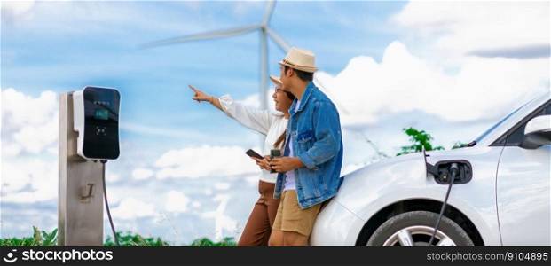 Concept of progressive happy young couple enjoying their time at windmill farm with electric vehicle. EV car driven by clean renewable energy from wind turbine generator for charging station.. Concept of progressive happy young couple at wind turbine with electric vehicle.