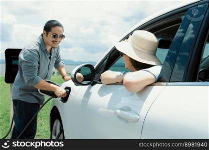 Concept of progressive happy young couple relax and enjoy natural green field and lake with electric vehicle. Electric vehicle driven by clean renewable. Refreshing environment from green energy.. Concept of progressive happy man and woman at green field with electric vehicle.