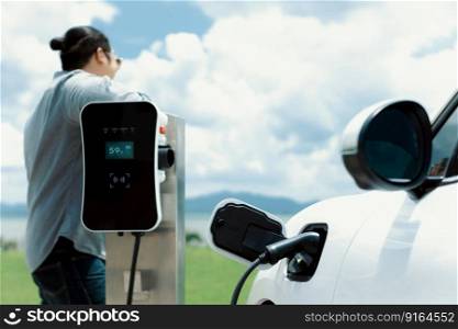 Concept of progressive man relax natural green field with electric vehicle. EV car driven by clean renewable plugged-in with charging point. Refreshing environment with cloudscape background.. Concept of progressive man at green field with plugged-in EV car and cloudscape.