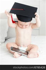 Concept of smart baby. Cute baby boy in graduation cap browsing internet on digital tablet