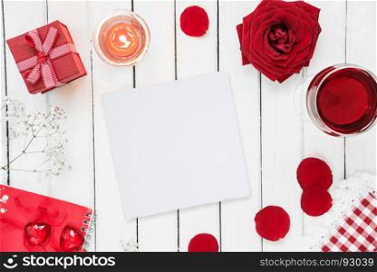 Concept of Valentine's Days holiday: festive table with glass of wine, red rose, gift box with bow and burning candle; top view, with copy-space on white paper card