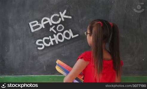 Concept on blackboard at school. Young people, student and pupil in classroom. Happy latina girl in class. Portrait of female child smiling, looking at camera, holding books. Back to school message