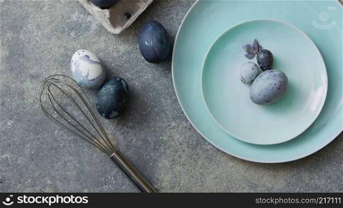 Concept preparation for Easter, blue-colored eggs on a plate with a whisk and dried flower on a concrete gray background, top view. Easter galactic eggs on plates with a whisk as a concept of cooking for Easter