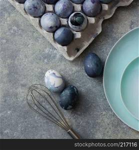 Concept preparation for Easter holiday, painted blue galactic eggs in a cardboard box with a whisk and blue plates on a concrete gray background, top view. Plate with painted eggs and whisk on a concrete background