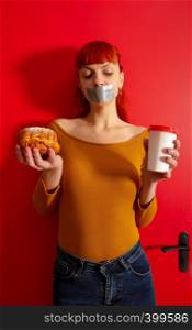 Conceptual image of a young red-haired girl on a diet sitting with her mouth taped with silver tape before appetizing pastries. diet lip scotch